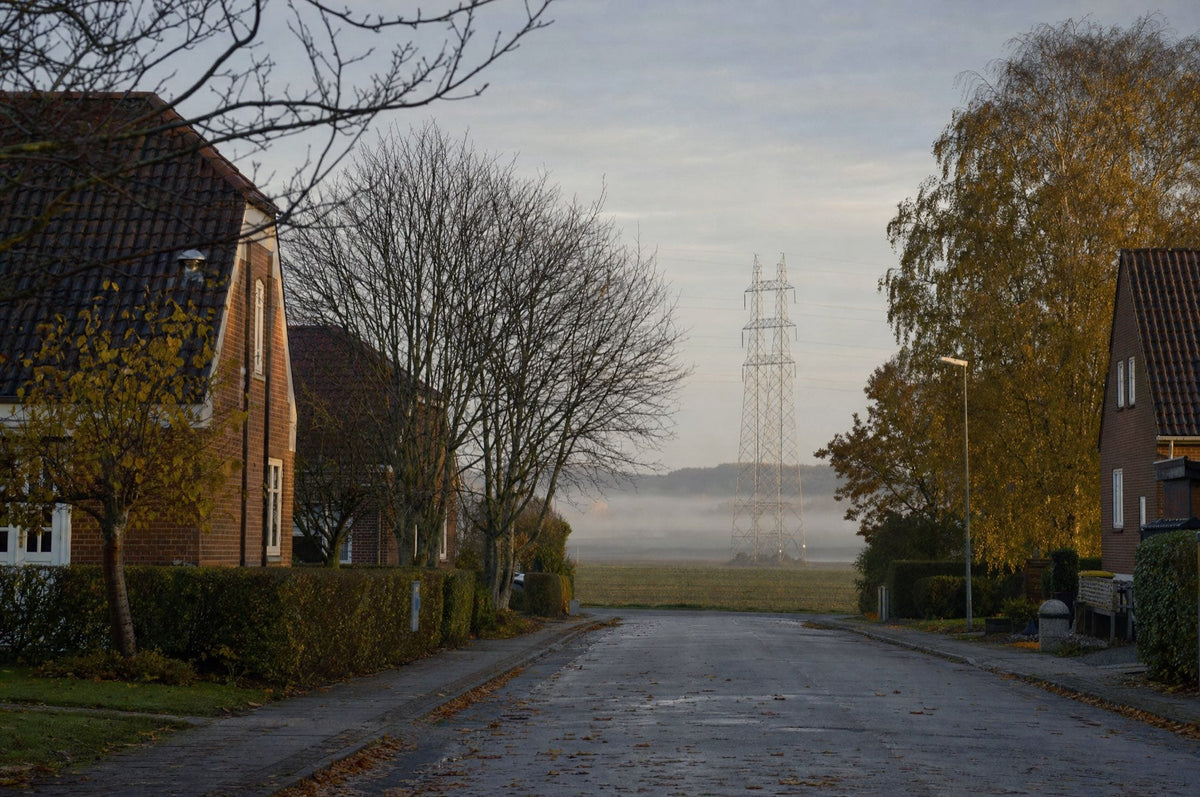Villavej i Danmark en tidlig morgen sidst i oktober, med røde murstenshuse, nøgne og gule efterårstræer og en højspændingsmast i morgendis på marken bagved. AI-genereret illustration.