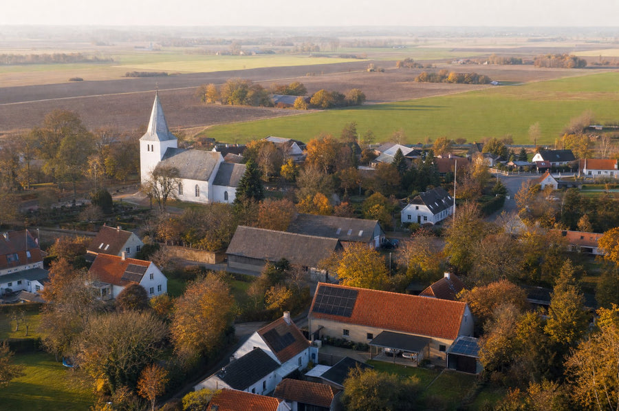 Luftfoto af en dansk landsby en sen eftermiddag i oktober: hvid kirke med spir, røde tegltage med enkelte solpaneler og træer i efterårsfarver. AI-illustration.