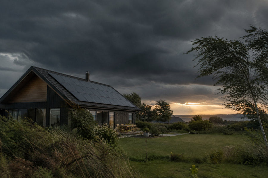 Et dansk parcelhus med solceller på taget under en mørk stormhimmel i vindstød, med bøjede træer og et lysbrud over horisonten, set fra haven.