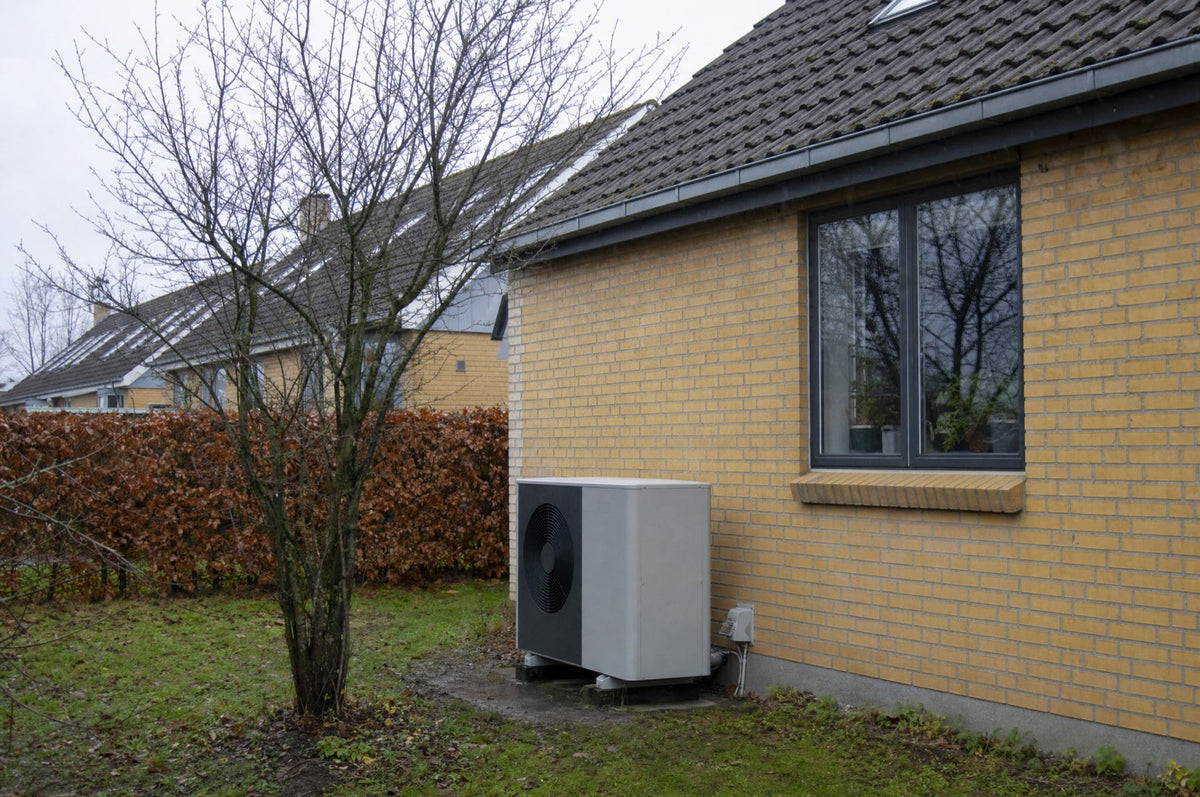 Gråt udendørsaggregat til en luft til vand varmepumpe på en lille sokkel ved gavlen af et gult murstenshus en gråvejrsdag i november, med bart træ og bøgehæk i haven